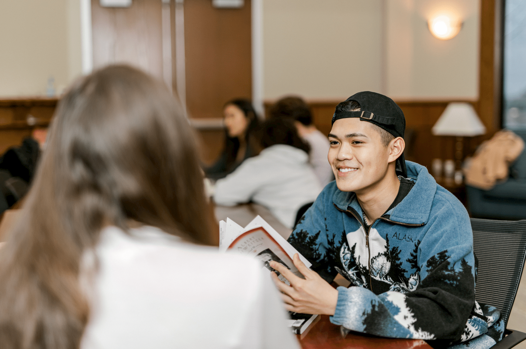 Sitting at a table, a student smiles as he looks up to talk to his peer