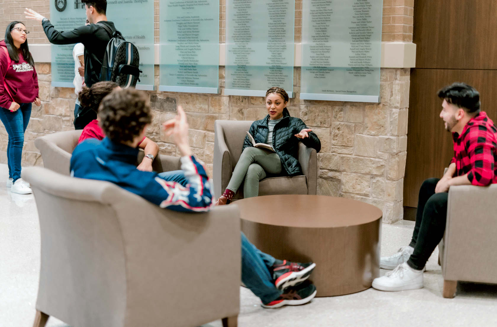 Four students sit in singular chairs and talk to each other across a brown coffee table.