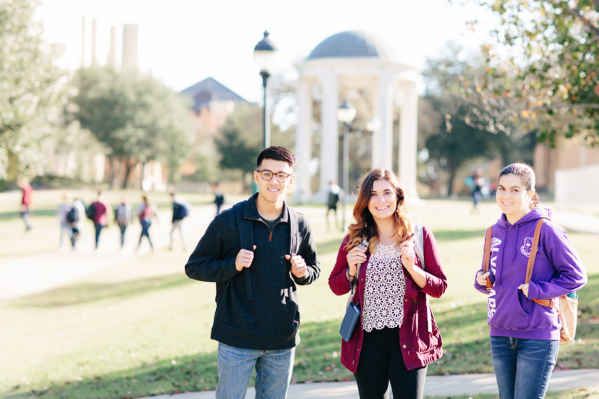 Three students stand togther smiling with their backpacks on infront of the rotunda while other students pass by