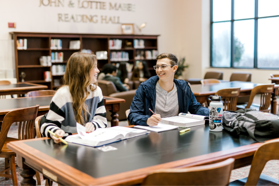 Two students sit together at the library looking at eachother smiling, they have pencilas and studying materials in front of them