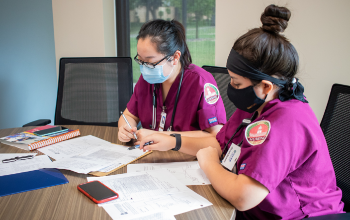 Two students, wearing maroon scrubs and masks, look down as they review their notes and study