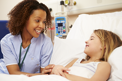 A nurse with light blue scrubs leans over as she talks to a child lying in a patient bed 