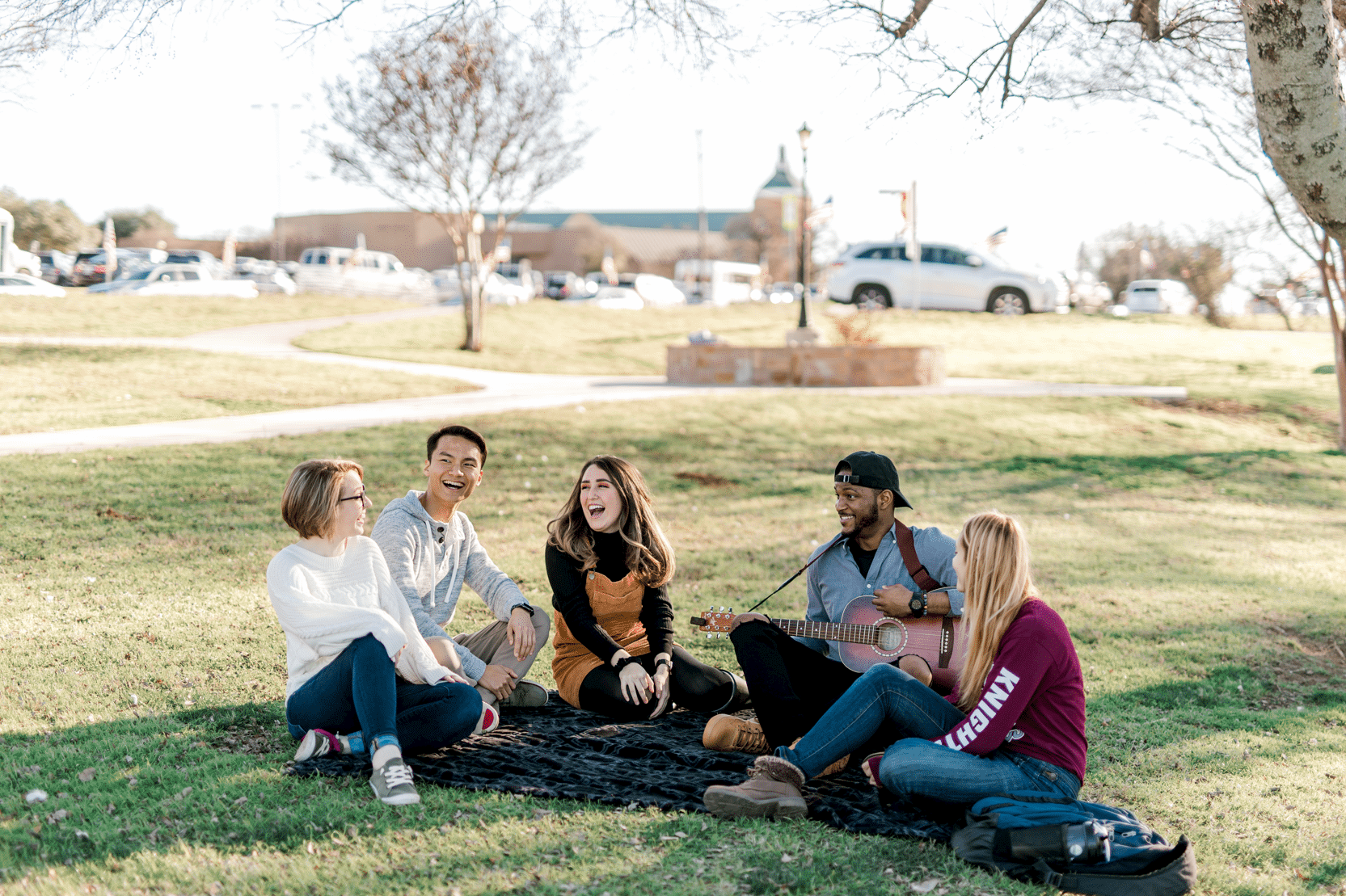 A group of students sit togther smiling and playing guitar on campus on the grass.