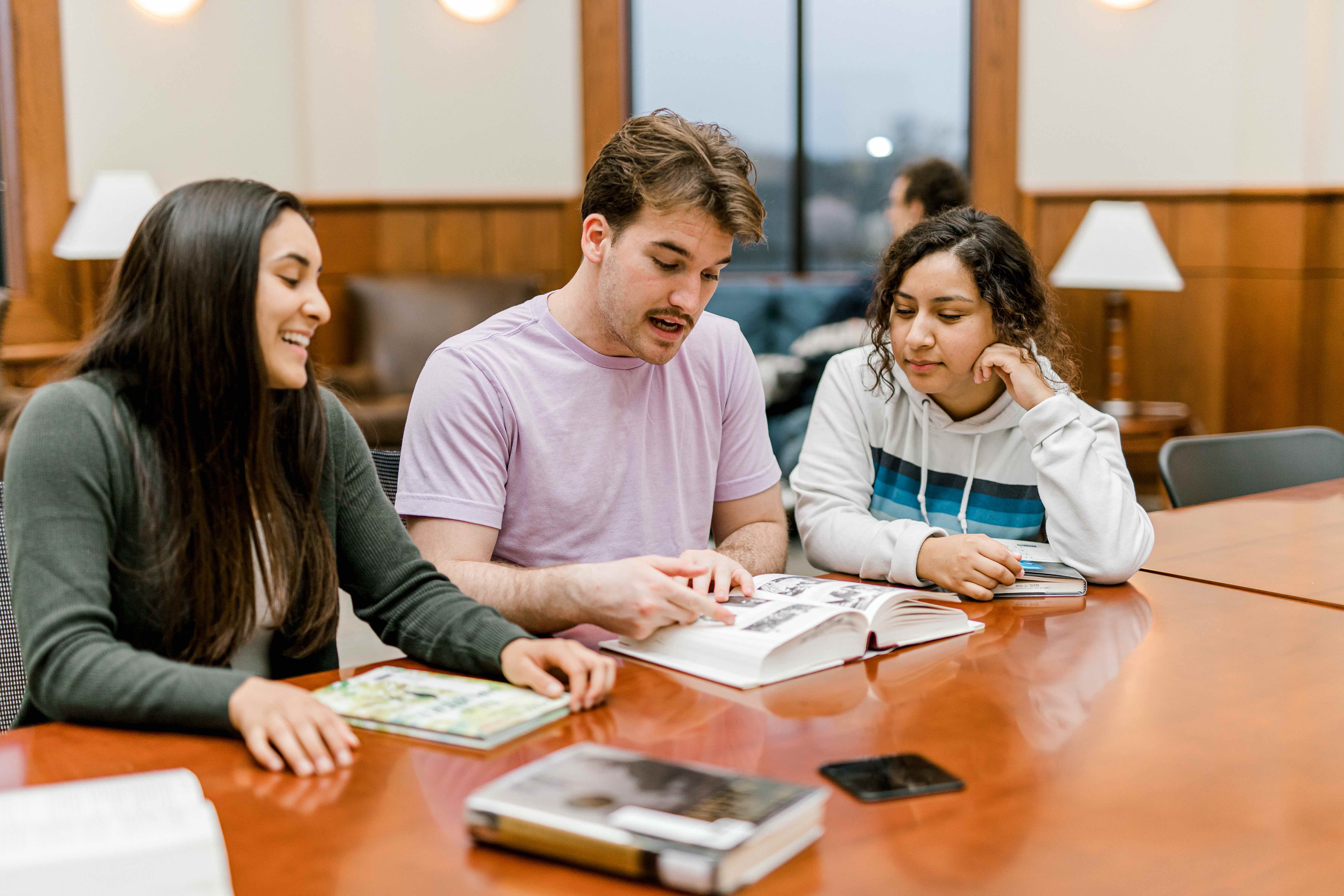 Three students look at a book as they sit at a rectangular table in the pechero student lounge