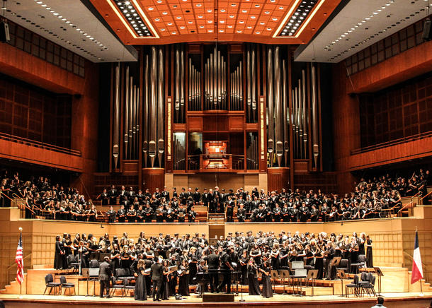 A mass choir with women in black gowns and men in all black suits stand in rows in front of a huge organ