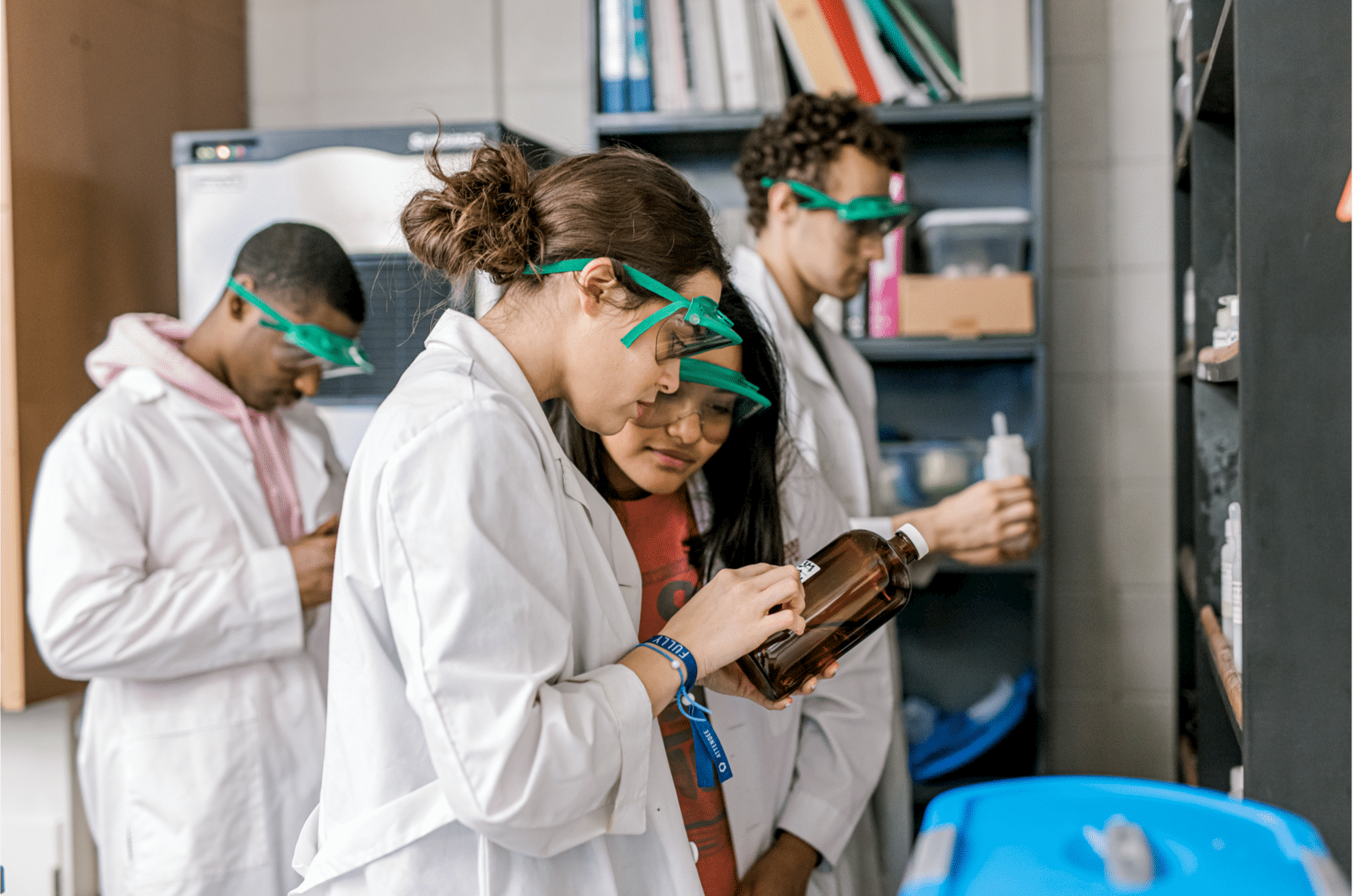 Wearing white lab coats and goggles, two female students lean in close as they read the label of a chemical bottle. Their peers can be seen in the back looking through other chemical bottles for their lab.