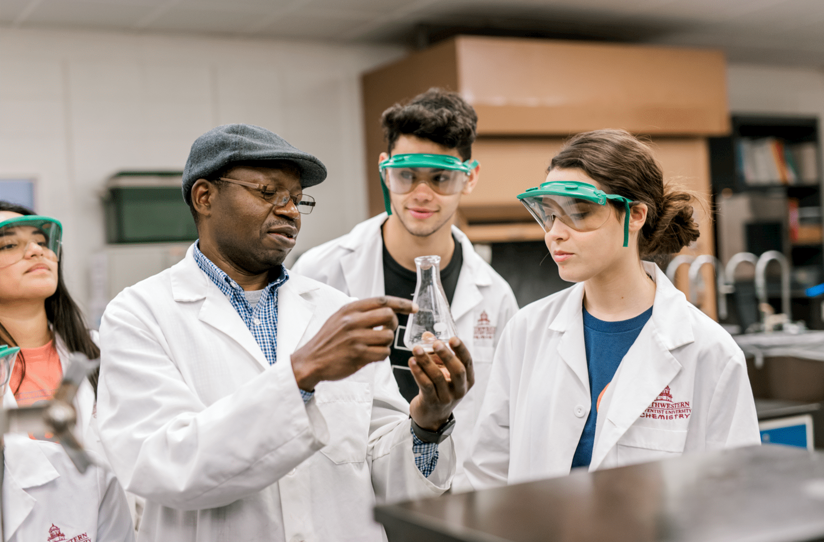 A professor holds up a flask as he points and explains something to his students. All of them are wearing lab coats while the students all wear goggles. 