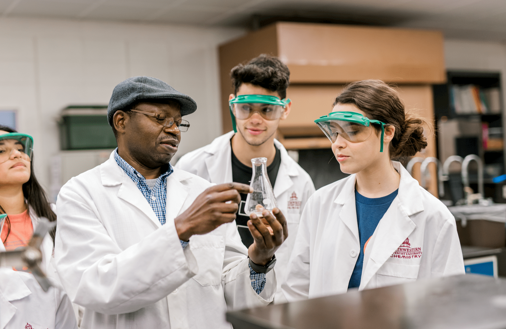 Two students wearing white lab coats and goggles listen to their professor who is holding and pointing to a flask 