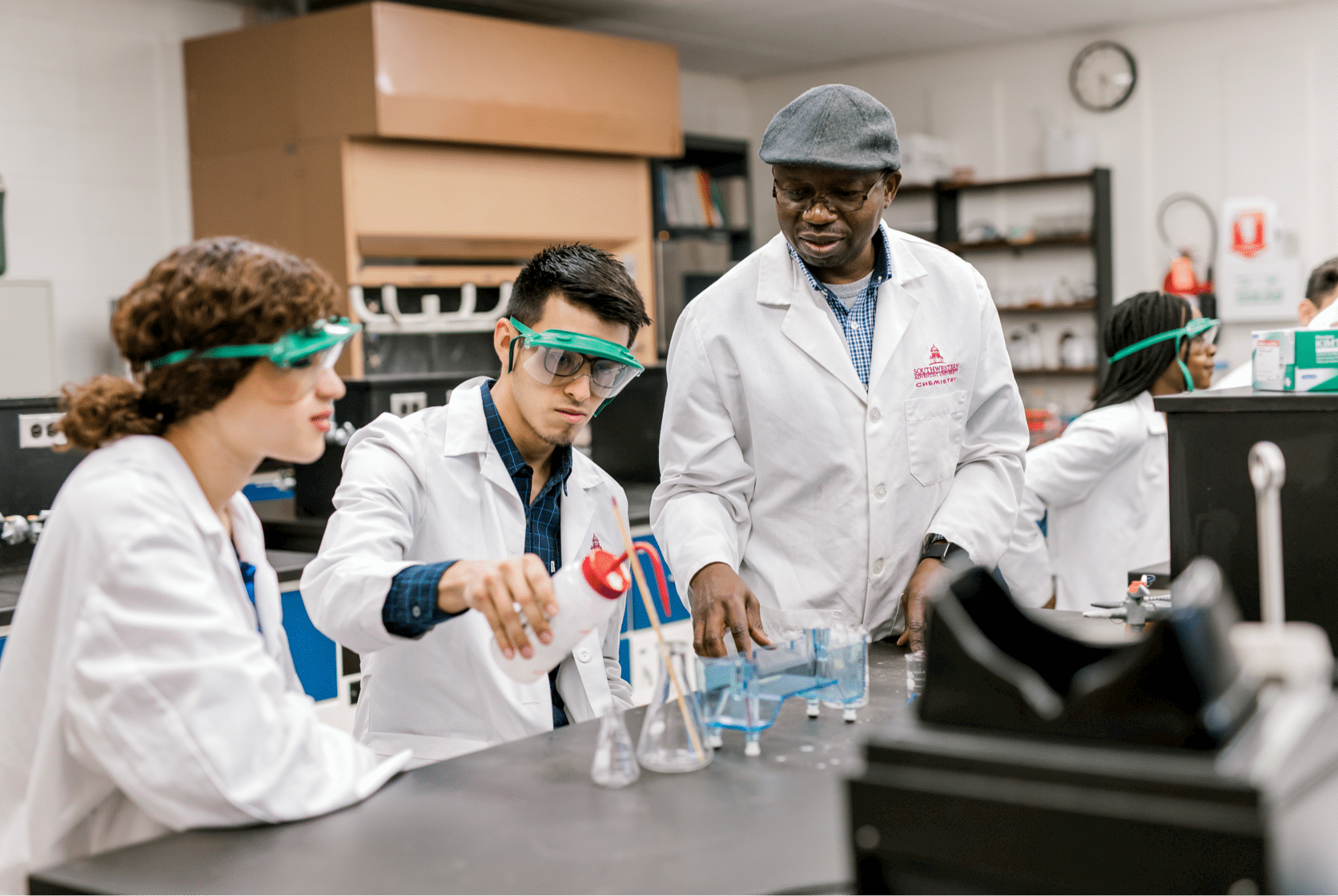 Two students, dressed in lab coats and googles, begin to conduct their lab with the help of their professor