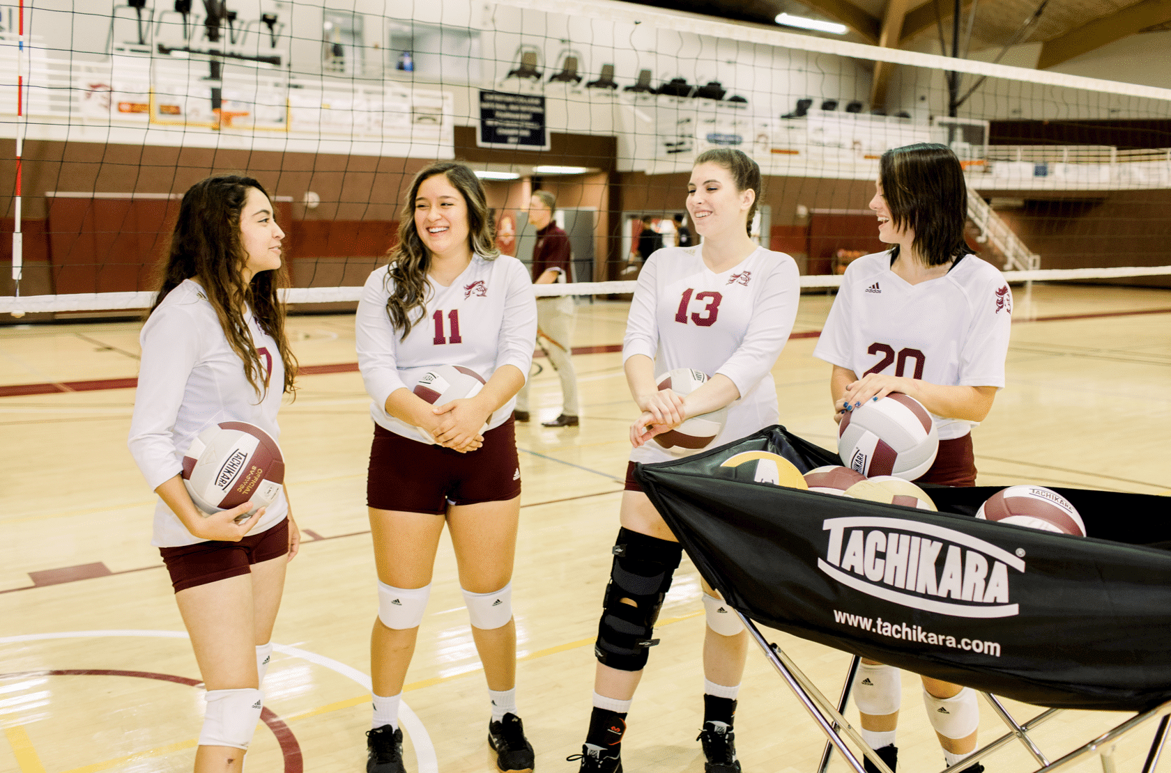 A group of volleyball players stand together talking, in uniform, each holds a volleyball