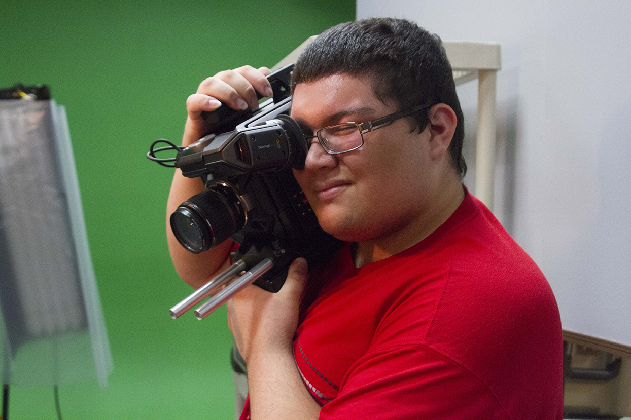 A young man holds a video camera on his should and squints to look through the lense