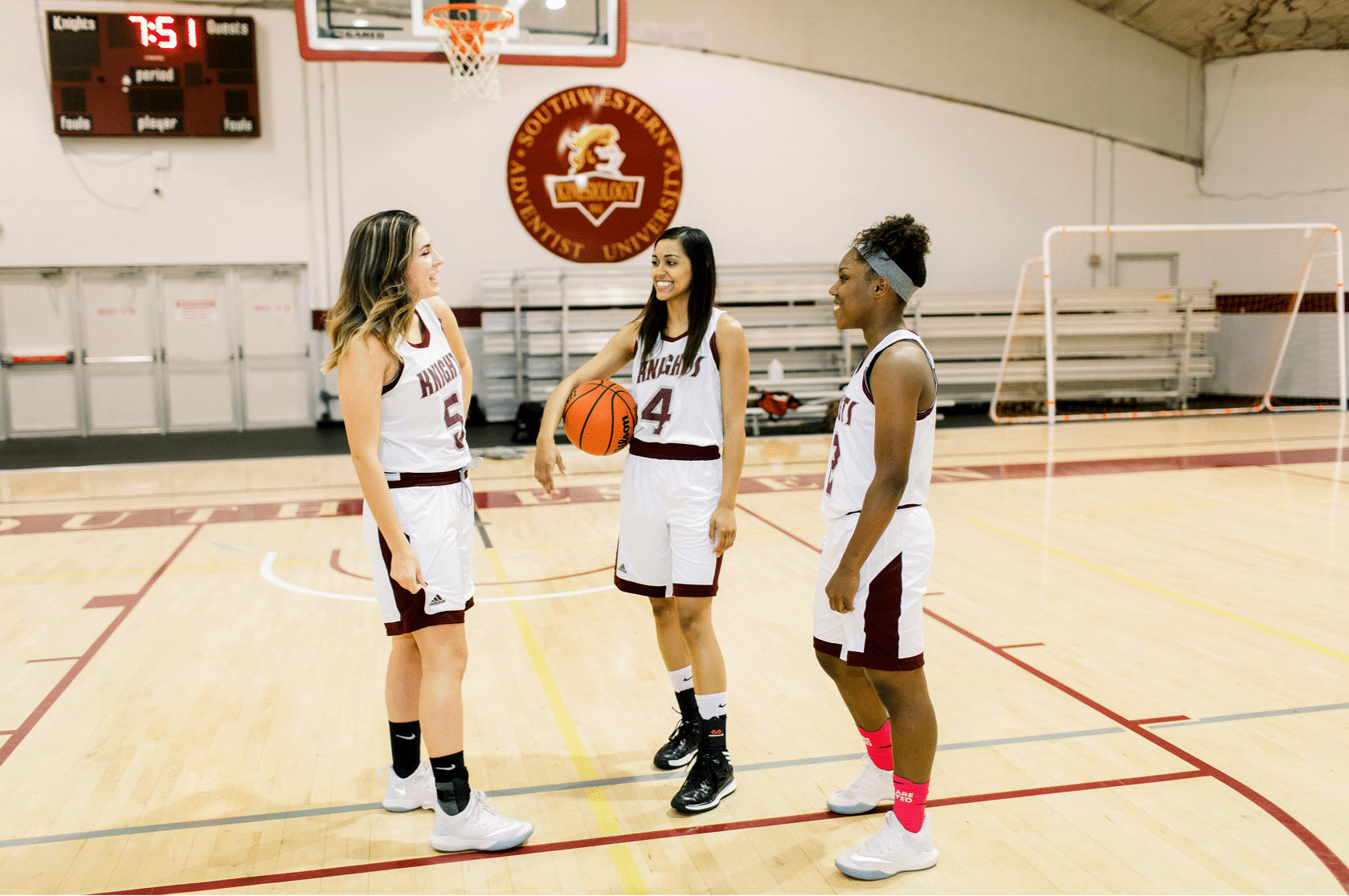 Three female basketball players in uniform stand talking together at the gymnasium, one holds a basketball