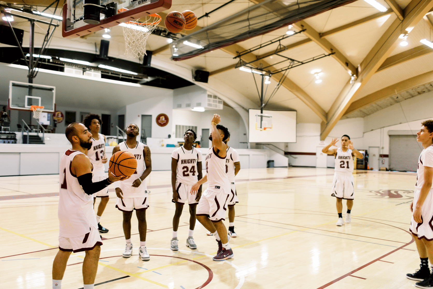 A group of male basketball players gather in the gymnasium wearing their uniforms as thye throw balls into the hoop and look around
