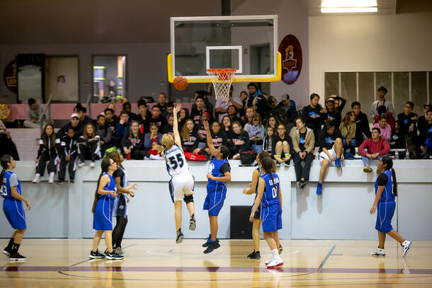 A girl, in a white basketball uniform, goes for a layup while the opponents, in blue uniforms stand back and watch