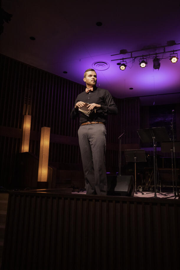 A youth pastor stand on top of a stage and holds his bible as he preaches and looks at the congregation.