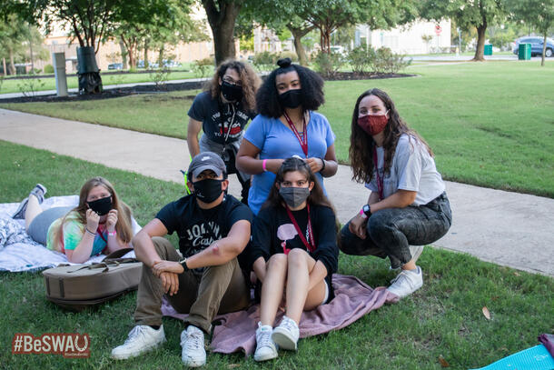 Sitting on blankets outside, six friends gather close and smile big under their face masks