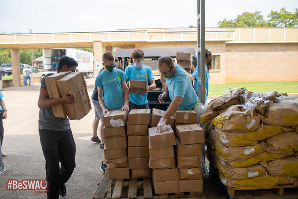 Four people wearing a blue LIFT t-shirt and masks carrying and stacking labeled cardboard boxes of food items 