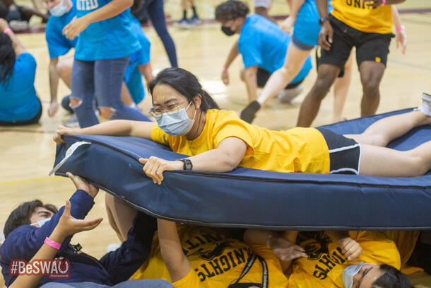 A student, wearing a yellow shirt to match her teammates, smiles as she lays on top of a matress that her teammates her holding and passing along.