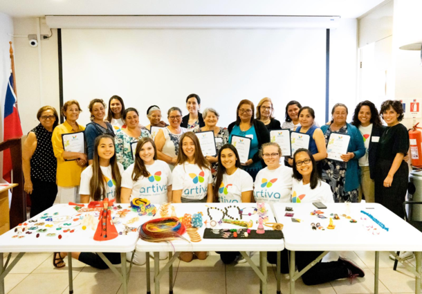 A group photo of women standsing with a certificate in hand, and the Enactus officers smiling while kneeling in front of a table of hand-made goods