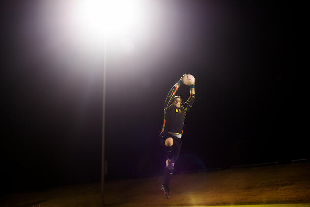 On the soccer field at night, the SWAU Men's goalie jumps up and blocks the ball