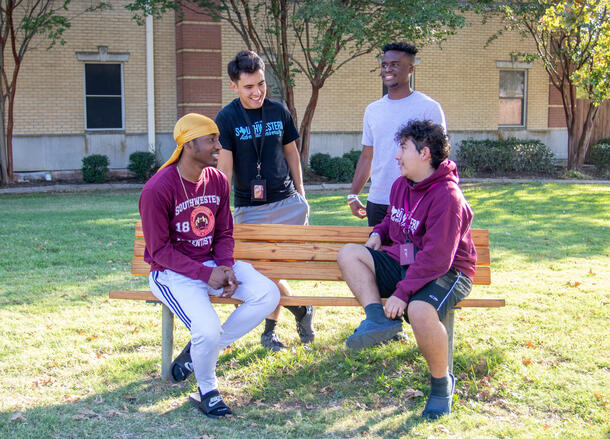 Four male students sit on and around a bench in front of the men's residence hall