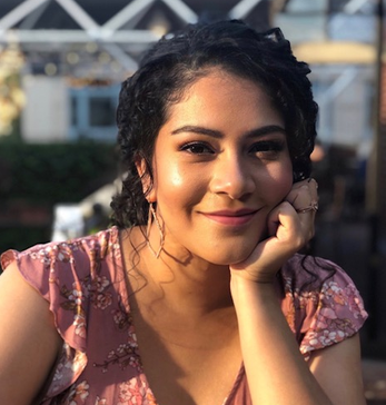 With her curly hair in a low pony tail, a young woman rests her face on her hand and gently smiles.