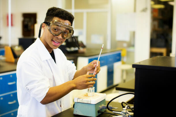 Wearing a white lab coat and goggles, a student looks up as he is holding a thermometer to take the temperature of his lab project