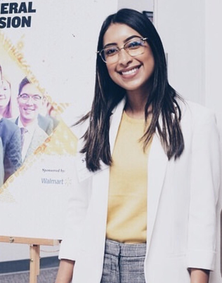 A woman wearing a white blazer, gray slacks and a yellow shirt, smiles as she stands in front of an Enactus Poster.
