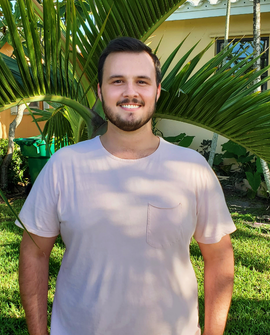 A young man with brown hair smiles as he stand in front of a small palm tree