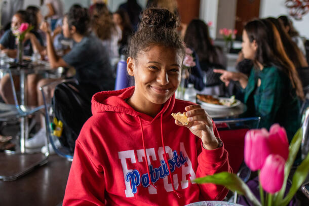 Girl in red hoodie poses smiling and holding half-eaten cookie with groups of girls at tables behind her