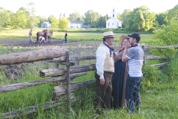 Outside where horses can be seen helping toil the ground, a man and woman stand outside a wooden fence, smiling as they talk to the director 