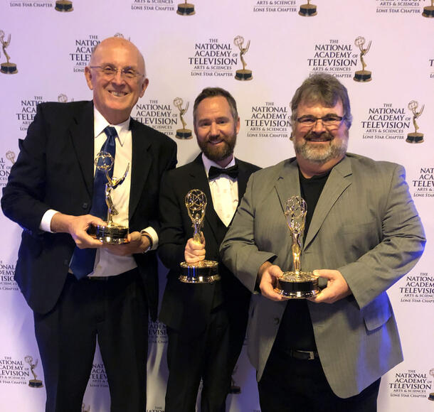 Standing in front of a backdrop, three men, dressed in suits, smile as they hold an award they received from the National Academy of Television.