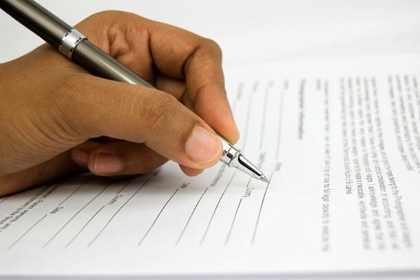 A hand holds a pen that hovers over a document, preparing to sign