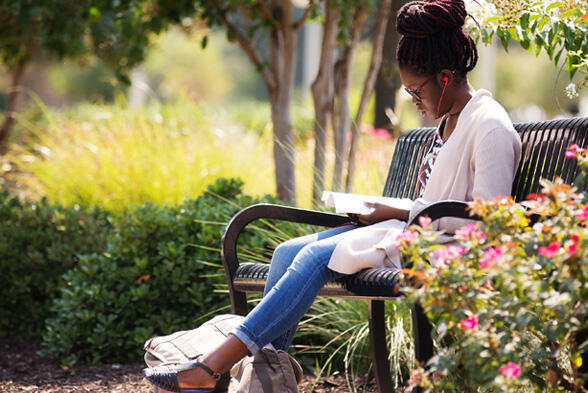 A student sits outside on a bench, reading a book while listening to music through her red headphones