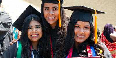 Three female students, dressed in their caps and gowns, smile as they hold their newly given diplomas 