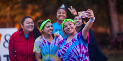 Five women gather around in bright shirts as they smile for a selfie 