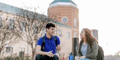 Two students sit together outside while looking at each other, talking and laughing 