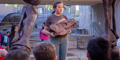 A dinosaur museum worker holds a dinosaur bone as she teaches the children around her