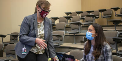 A professor wearing a gray blazer and a maroon masks stands beside a seated student to talk