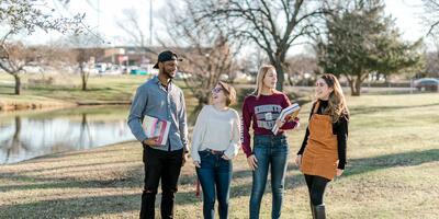 Four students, two with books in hand, laugh as they talk and stand in front of the duck pond.