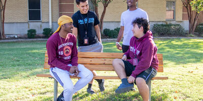 Four male students sit on and around a bench in front of the men's residence hall