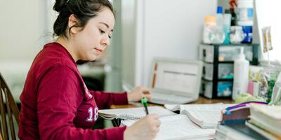 A student, dressed in a maroon long-sleeved shirt and with her hair in a bun, sits in front of a table as she completes her homework 