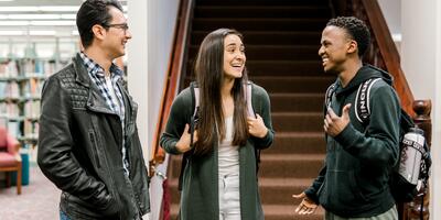 Three students stand in front of the carpeted-library stairs and laugh 