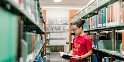 A male student with his hair tied up stands in between the library shelves as he reads through a book