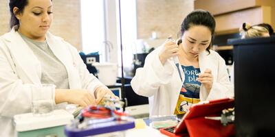Two female students wearing lab coats, focus on their expirement 