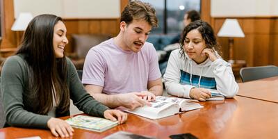 Three students look at a book as they sit at a rectangular table in the pechero student lounge
