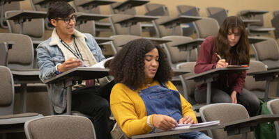 Three students sit down in a large classroom and begin to take notes of the lecture
