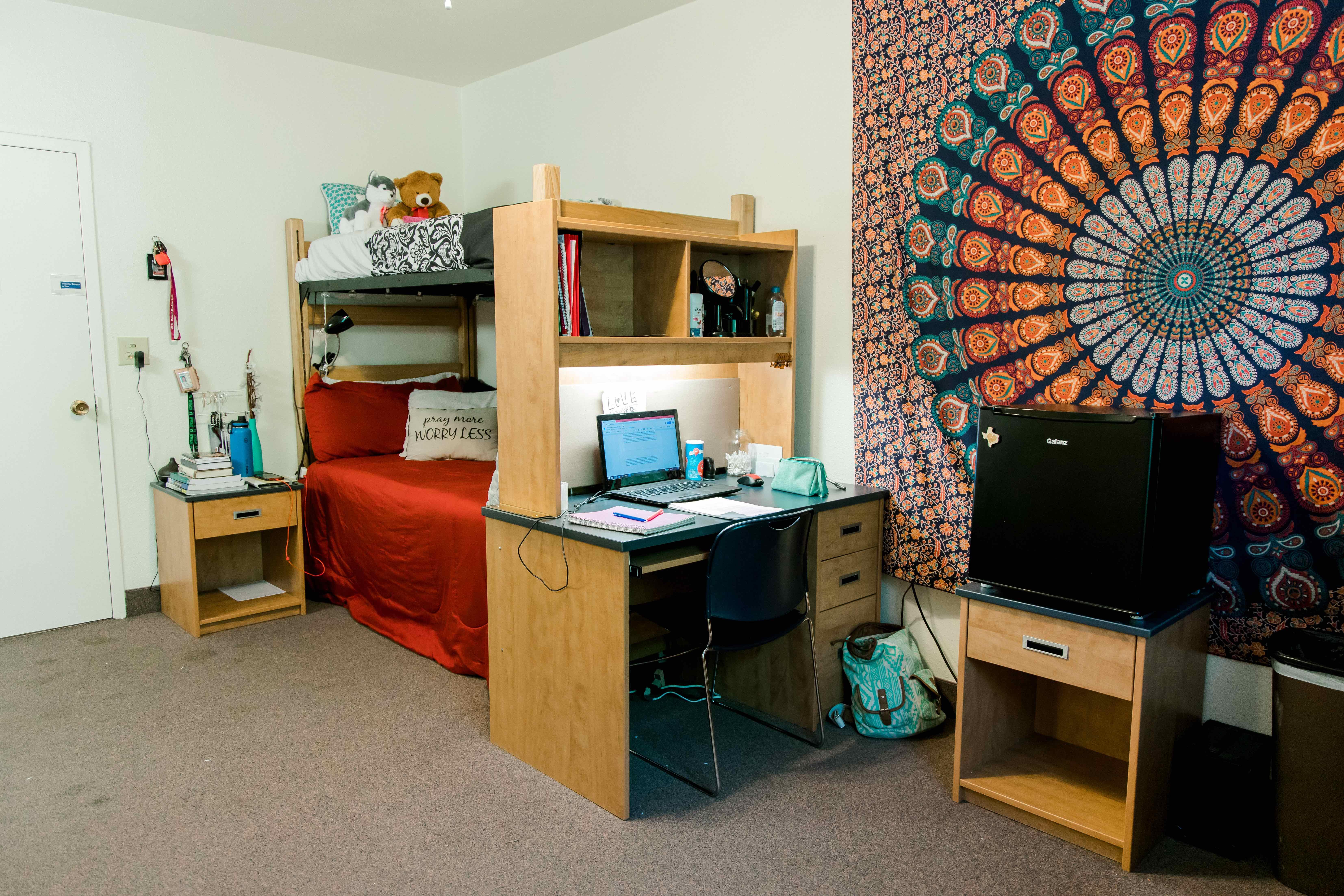 Inside the room of a dorm, a bunkbed is seen with desks on the otherside. On the wall is a large cloth with vibrant colors.