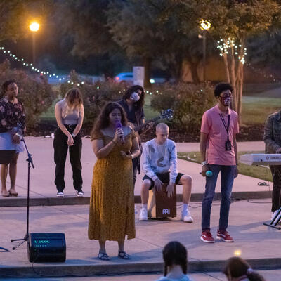 A group of students, a few standing by with instruments, stand outside with their eyes closed as they bow their heads to pray.