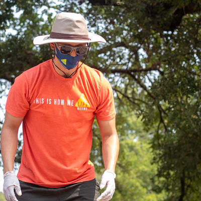 A male student, wearing an orange t-shirt and a bucket hat, looks down as he prepares to pick up something with gloves.